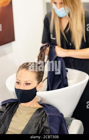 Donna parrucchiera che lava la testa di un cliente in un salone, protetto da una maschera Foto Stock