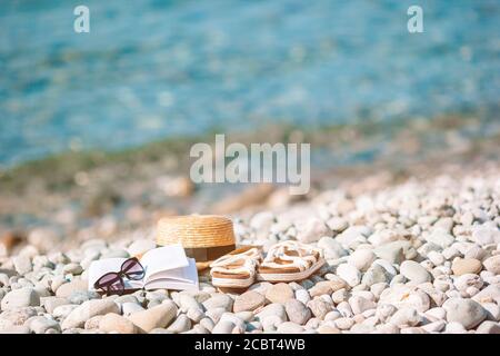 Cappello da spiaggia su libro aperto con crema solare e scarpe sopra spiaggia di ciottoli Foto Stock