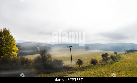 Paesaggio panoramico autunnale con linea elettrica e nebbia sopra prati e colline boscose. Panorama malinconico di prateria con vecchi tralicci elettrici. Foto Stock