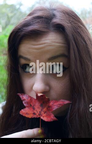 Una giovane donna con occhio pesante compongono e lungo capelli brunette che tengono una foglia rossa caduta acer a lei labbra Foto Stock