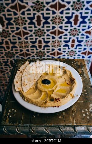 Hummus di ceci, patatine di pita, paprika e prezzemolo. Su un piccolo tavolo rustico in legno e tipiche piastrelle arabe sullo sfondo Foto Stock