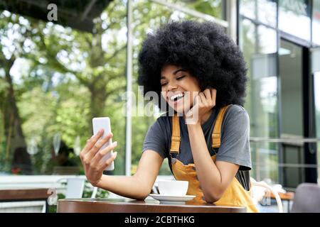 Felice ragazza teen africana che fa la videochiamata sul telefono siede al tavolo esterno del caffè. Foto Stock