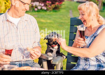 Felice coppia anziana che alimenta cane con pizza durante la cena cortile Foto Stock