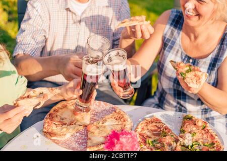 Immagine ritagliata di persone felici che mangiano pizza e tostano con birra all'aperto al tramonto - concetto familiare (attenzione ai bicchieri) Foto Stock