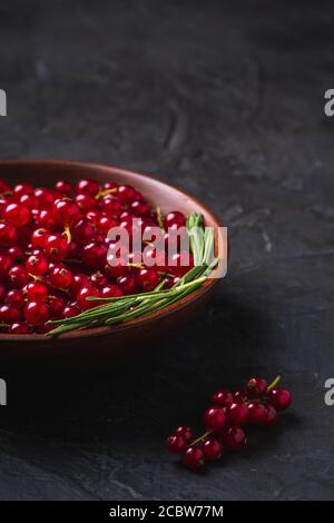 Bacche di ribes rosso dolce fresche con foglie di rosmarino in ciotola di legno, fondo scuro a trama, vista angolare Foto Stock