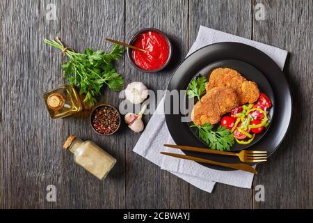 costolette di agnello impanate servite con pomodoro, cipolla rossa, insalata di pepe verde su un piatto nero su un tavolo di legno con ingredienti sullo sfondo, paesaggio Foto Stock