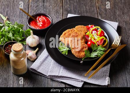 costolette di agnello impanate al forno servite con pomodoro, cipolla rossa, insalata di pepe verde su un piatto nero su un tavolo di legno, vista orizzontale, primo piano Foto Stock