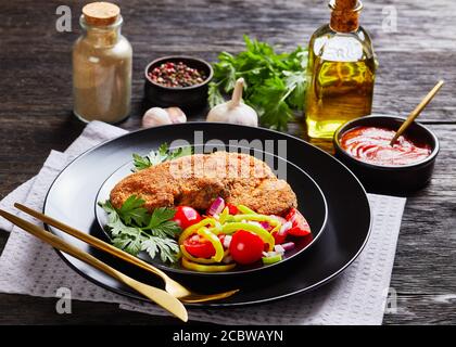 costolette di agnello impanate servite con pomodoro, cipolla rossa, insalata di pepe verde su un piatto nero su un tavolo di legno con ingredienti sullo sfondo, paesaggio Foto Stock