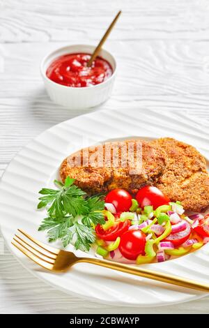 costolette di agnello impanate servite con pomodoro, cipolla rossa, insalata di pepe verde su un piatto bianco su un tavolo di legno, primo piano, vista verticale dall'alto Foto Stock