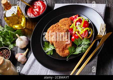 costolette di agnello impanate servite con pomodoro, cipolla rossa, insalata di pepe verde su un piatto nero su un tavolo di legno, vista panoramica dall'alto Foto Stock