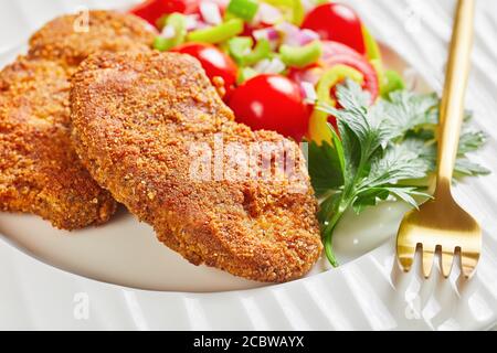 costolette di agnello impanate servite con pomodoro, cipolla rossa, insalata di pepe verde su un piatto bianco su un tavolo di legno, primo piano Foto Stock