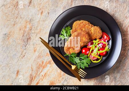 costolette di agnello impanate al forno servite con pomodoro, cipolla rossa, insalata di pepe verde su un piatto nero su un tavolo di pietra Foto Stock