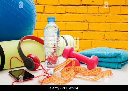 Palla fitness, manubri, tappetino, smartphone e auricolari su sfondo giallo in mattoni Foto Stock