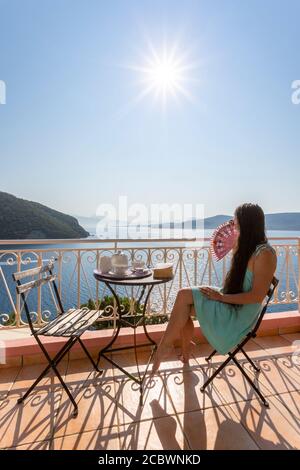 Una ragazza in abito si siede su una villa balcone con caffè al mattino e si affaccia su una vista costiera, Desimi, Lefkada, Isole IONIE, Grecia Foto Stock