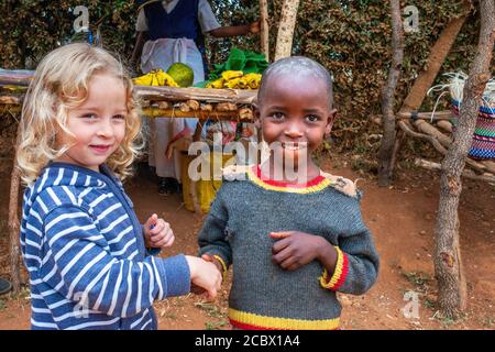 Ragazza bionda europea che dà la mano e la condivisione con un ragazzo nero nella scuola primaria e seconda in Un piccolo villaggio vicino alla città di Kitui nel Foto Stock