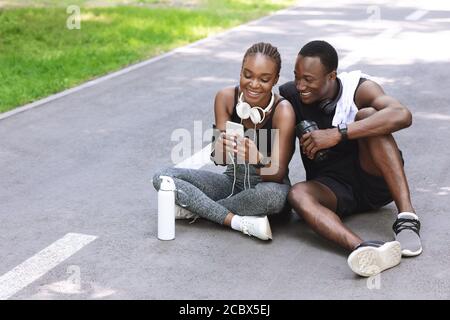 Una coppia gioiosa nera che si rilassa dopo un allenamento in fitness all'aperto, utilizzando uno smartphone Foto Stock
