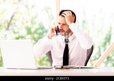 Un'immagine tagliata di un uomo d'affari stressato con la mano sulla fronte che lavora sul computer portatile e parla sul telefono cellulare mentre si siede in ufficio. Foto Stock