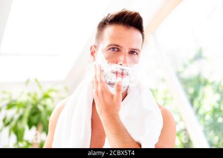 Foto di un bell'uomo con un asciugamano intorno al collo utilizzando la schiuma da barba prima di radersi mentre si trova davanti allo specchio del bagno. Foto Stock