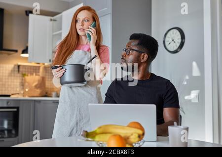 donna parlare al telefono mentre il marito lavora sul computer portatile a casa, nero ragazzo guardare donna di conversazione, lei tiene la pentola in mani Foto Stock
