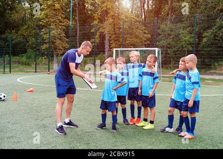 Il giovane allenatore insegna ai bambini la strategia di giocare sul campo di calcio. Foto Stock