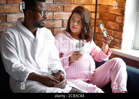 donna caucasica incinta con uomo nero. mix di razza concetto di relazione. si siedono sul divano a casa, tempo libero nei fine settimana Foto Stock