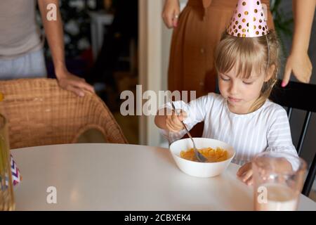 la bambina si siede facendo colazione con cappello da festa, la ragazza si siede da sola in cucina. festa, bambini, compleanno, concetto di colazione Foto Stock
