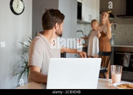 l'uomo che lavora voltò la testa indietro, guarda la moglie e la figlia in cucina. freelance, lavoro da casa concetto Foto Stock