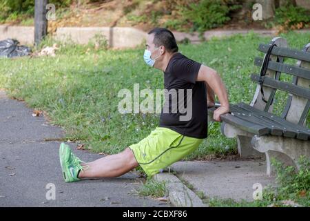 Un uomo americano asiatico che indossa una maschera chirurgica fa esercizi di immersione panca in un parco a Flushing, Queens, New York City. Foto Stock