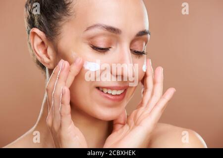 Giovane donna allegra con sorriso toothy applicando crema facciale ringiovanente Foto Stock