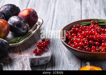 Frutta fresca di prugne dolce intera e tagliata in piatto con foglie di rosmarino sul vecchio tagliere con bacche di ribes rosso in una ciotola di legno Foto Stock
