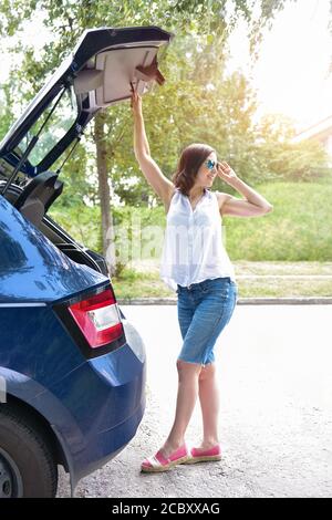 Donna caucasica sorridente in camicetta bianca e pantaloncini in denim mettendo le sue borse di roba nel bagagliaio dell'automobile Foto Stock