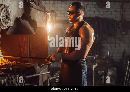 uomo bearded in buona forma fisica con i muscoli forti che scaldano il ferro per la produzione, indossando grembiule di cuoio e occhiali protettivi isolati in w Foto Stock