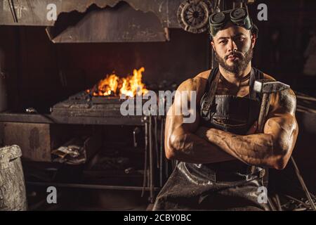 Ritratto di un giovane metalworker serio e sicuro in un grembiule nero che guarda la macchina fotografica, lavora in officina Foto Stock