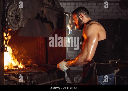 vista laterale su uomo sinewy bearded con corpo muscolare riscaldamento ferro nel fuoco, indossando grembiule in pelle isolato in officina scura Foto Stock