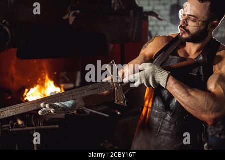 giovane uomo di forger bearded che studia metallo fatto a mano in officina vicino al forno, indossando grembiule di cuoio marrone, guanti e occhiali protettivi Foto Stock