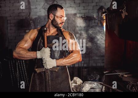 muscoloso fabbro maschio che tiene in mano metallo fatto a mano e guardando il lato, uomo caucasico bearded che indossa guanti protettivi per lavoro e occhiali per Foto Stock