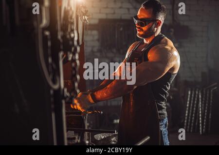 vista laterale su uomo sinewy bearded con corpo muscolare riscaldamento ferro nel fuoco, indossando grembiule in pelle isolato in officina scura, in occhiali protettivi Foto Stock