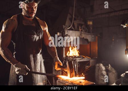 l'uomo giovane con la bearded in uniforme di cuoio riscalda il metallo sopra isolamento antincendio in officina Foto Stock