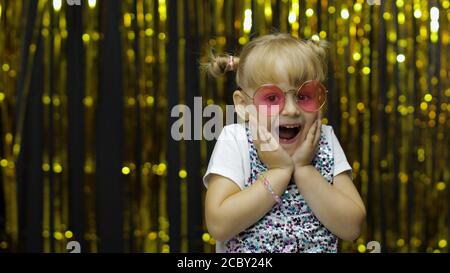 Bambini che ballano, show stupement, imbrogliare intorno, sorridere, guardare sorpreso scioccato. Piccolo divertimento biondo bambino teenager ragazza 4-5 anni in occhiali da sole che posa isolato su sfondo con tenda di lamina Foto Stock