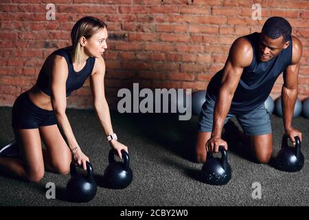 Giovane atleta caucasica facendo affondare su kettlebells con il aiuto del personal trainer durante l'allenamento crossfit del corpo Foto Stock