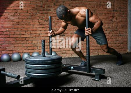 Bodybuilder maschile skinned scuro che esegue peso pesante slitta spingere l'esercizio in cross fit palestra. Foto Stock