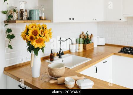 Piatti e utensili sul tavolo da cucina, pronti per cucinare. Bianco semplice cucina moderna in stile scandinavo, dettagli cucina, tavolo in legno, girasoli bouq Foto Stock