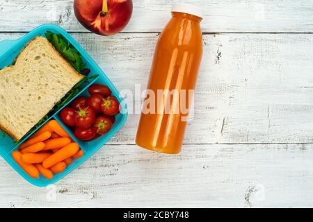 Pranzo scolastico - scatola di plastica con sandwich, pomodori, carote in sezioni con succo d'arancia in bottiglia su sfondo di legno bianco. Foto Stock