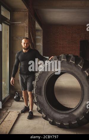 CrossFit tecnica dei pneumatici. Potente bodybuilder spiega le regole e ordine di formazione con improvvisato powerlifting elemento, in piedi accanto a un enorme pneumatico in fi Foto Stock