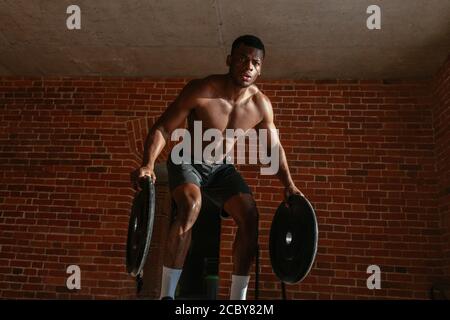 Potente forte combattente africano boxer pratica salti ponderata con barbell piastre su e su grandi giacente carrello pneumatico in crossfit studio contro il mattone Foto Stock