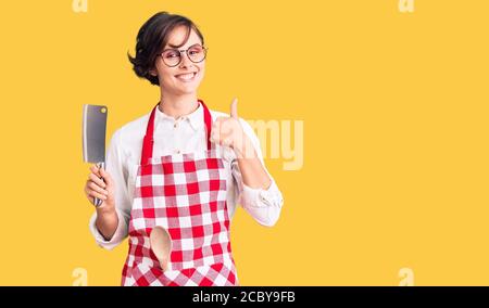 Bella giovane donna con capelli corti indossando grembiule professionale che tiene il coltello sorridente felice e positivo, pollice su facendo eccellente e approvazione Foto Stock