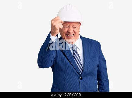 Uomo anziano bello grigio-capelli che indossa vestito e architetto hardhat arrabbiato e folle alzando pugno frustrato e furioso mentre gridando con rabbia. Rabbia A. Foto Stock