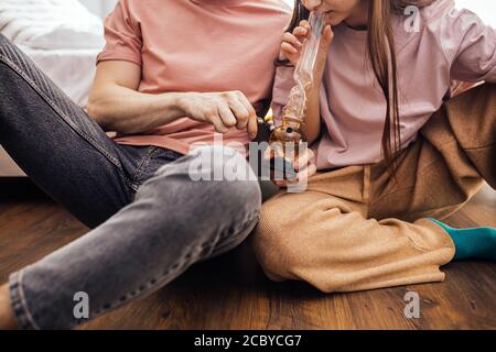 giovane uomo e donna caucasica che usa droghe a casa, coppia soffre di dipendenza da droga insieme, usa l'erbaccia di cannabis con il bong. all'interno, siediti sul pavimento Foto Stock