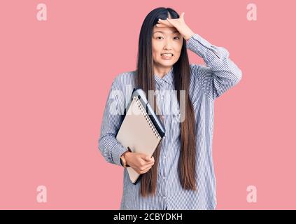 Giovane bella donna cinese che tiene taccuino stressato e frustrato con la mano sulla testa, sorpresa e faccia arrabbiata Foto Stock