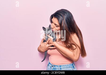 Giovane bella donna latina che puzzava felice. In piedi con sorriso sulla faccia che tiene e baciare adorabile gatto su isolato sfondo rosa Foto Stock